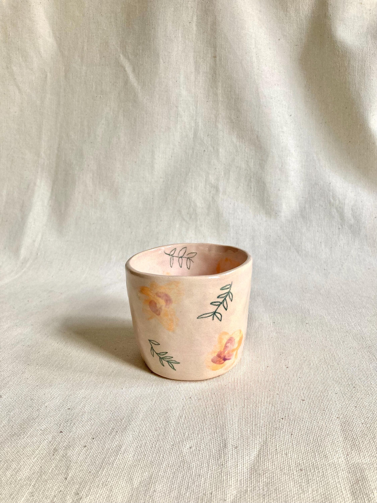 Peut inclure: Une petite tasse en céramique faite à la main, avec une base rose clair. La tasse est décorée de motifs floraux et de feuilles de style aquarelle dans des tons de jaune, d'orange et de vert. La tasse a une finition lisse et mate, idéale pour petits objets.