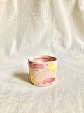 Peut inclure: Une petite tasse en céramique peinte à la main, avec une base rose clair. La tasse est décorée de coups de pinceau jaunes et rouges et de petits points noirs. Le bord intérieur est rose plus foncé. La tasse repose sur un tissu de couleur neutre.