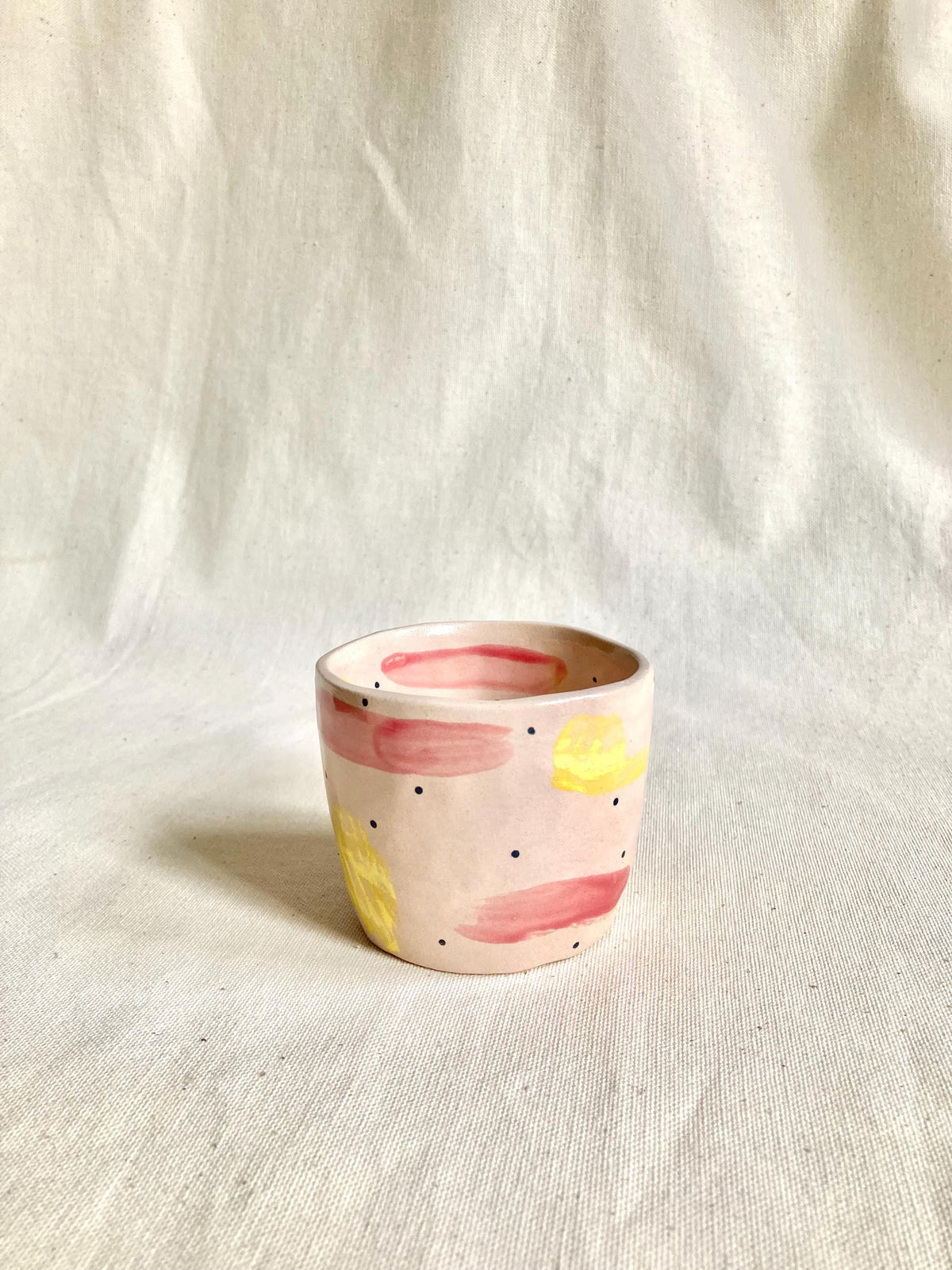 Peut inclure: Une petite tasse en céramique peinte à la main, avec une base rose clair. La tasse est décorée de coups de pinceau jaunes et rouges et de petits points noirs. Le bord intérieur est rose plus foncé. La tasse repose sur un tissu de couleur neutre.