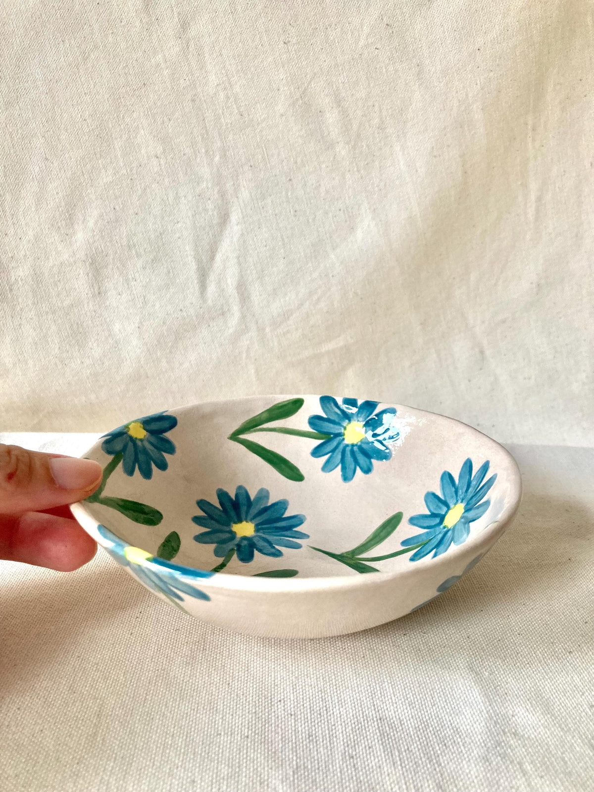 Peut inclure: Un petit bol en céramique blanc cassé décoré de fleurs de marguerite bleues et de feuilles vertes. Les fleurs ont des centres jaunes. Le bol est peint à la main et a une forme légèrement irrégulière, suggérant qu'il est fait à la main.