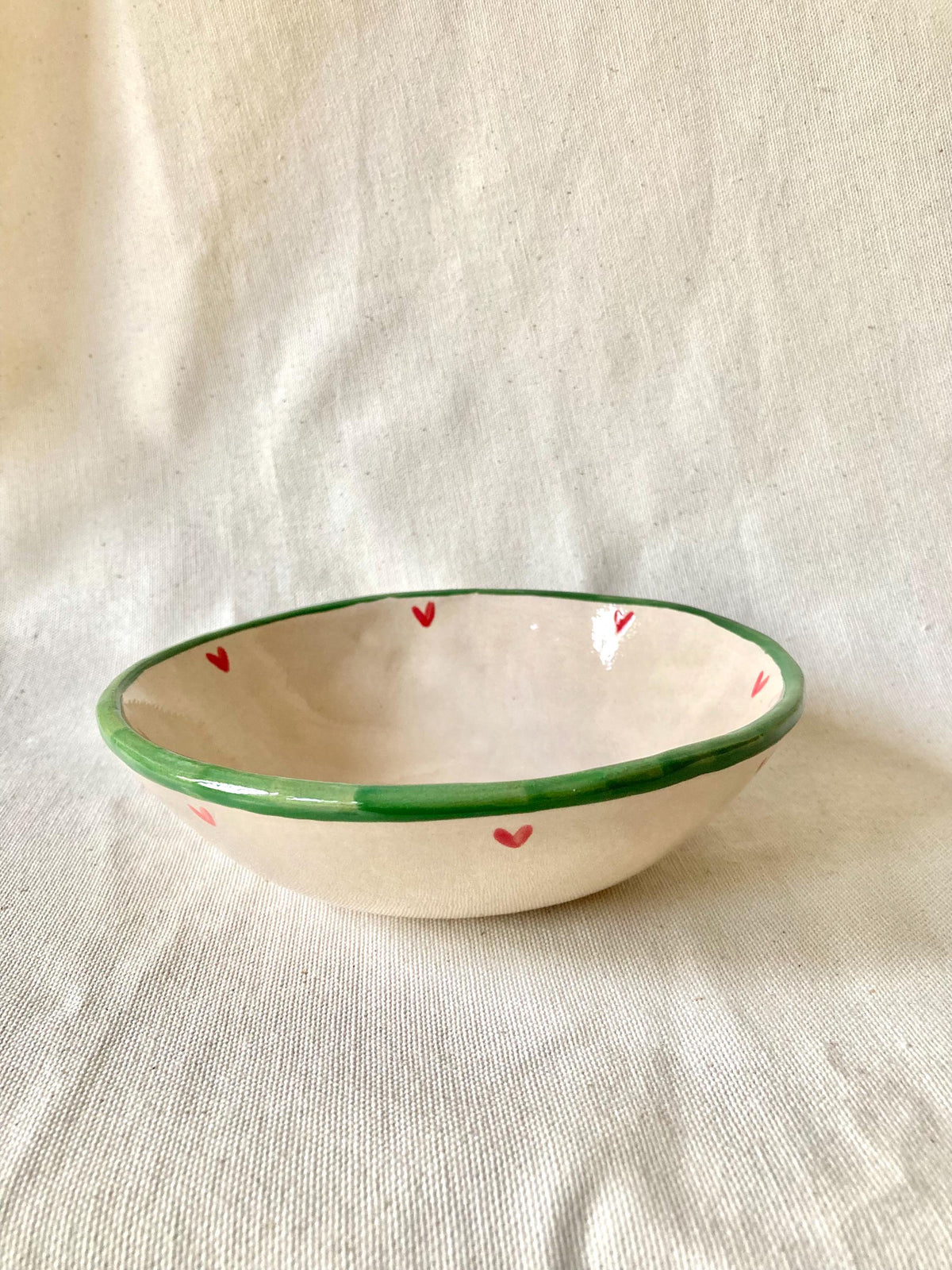 Peut inclure: Un bol en céramique fait à la main, intérieur crème et bord vert. De petits cœurs rouges ornent l'extérieur. Le bol est posé sur un fond en tissu neutre.