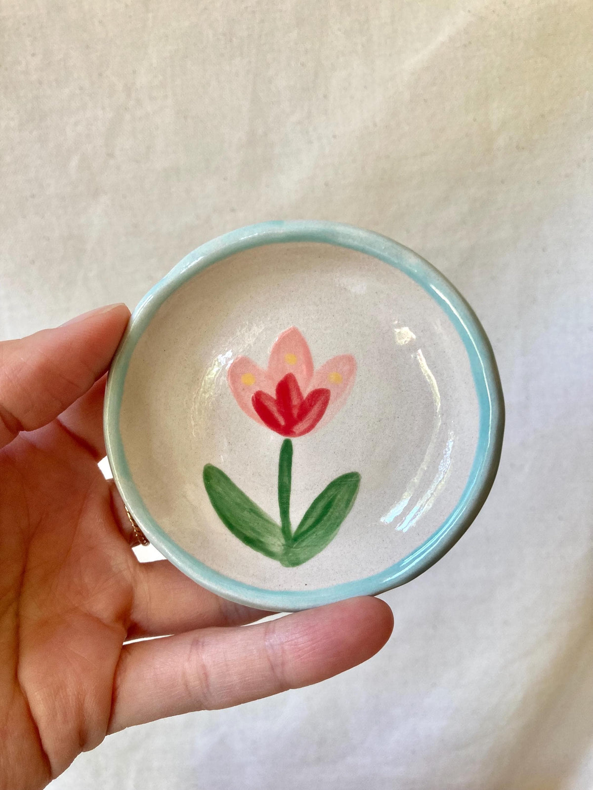 Peut inclure: Petit plat rond en céramique avec un motif de tulipe peint à la main. La tulipe présente une fleur rouge, une tige et des feuilles vertes et des accents jaunes. Le bord est souligné en bleu clair. Le plat mesure environ 10 cm de diamètre.
