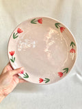Peut inclure: Une assiette ronde en céramique rose clair avec un motif floral. L'assiette présente une bordure de fleurs rouges et de feuilles vertes. L'assiette est tenue par une main sur un fond blanc.