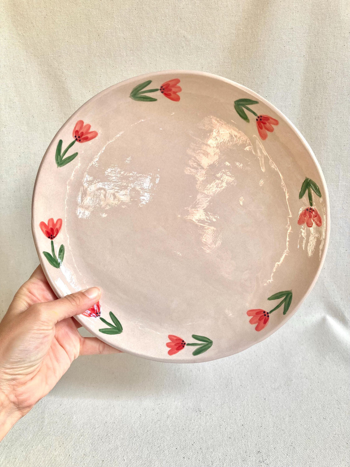 Peut inclure: Une assiette ronde en céramique rose clair avec un motif floral. L'assiette présente une bordure de fleurs rouges et de feuilles vertes. L'assiette est tenue par une main sur un fond blanc.