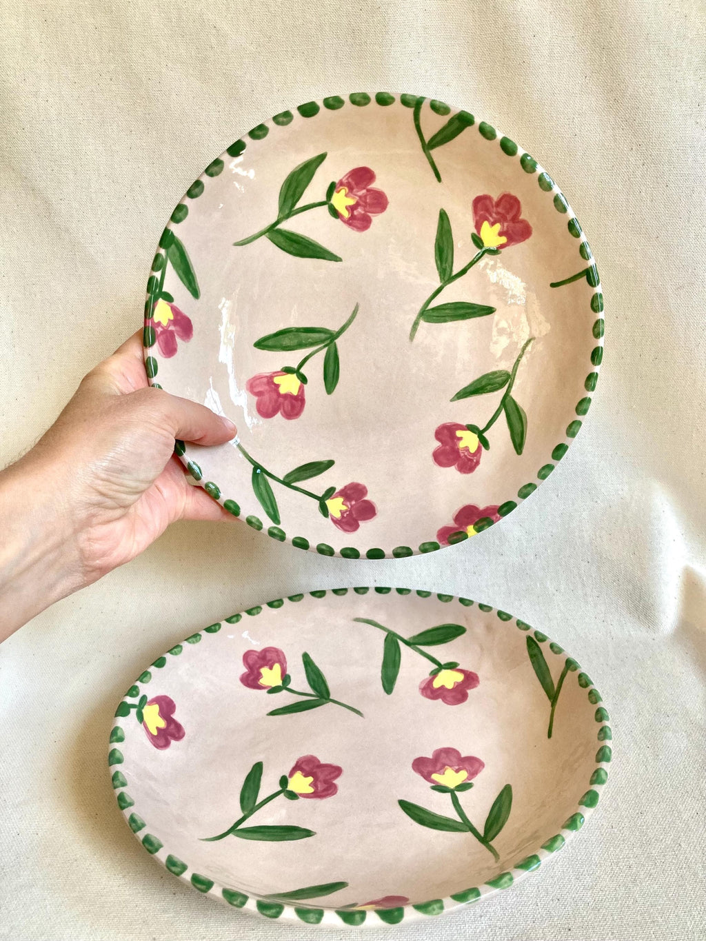 Peut inclure: Deux assiettes en céramique avec un fond rose et un motif floral. Les fleurs sont rouges avec des centres jaunes et des tiges et feuilles vertes. Les bords sont décorés d'une bordure verte pointillée. Une assiette est ronde, l'autre ovale.