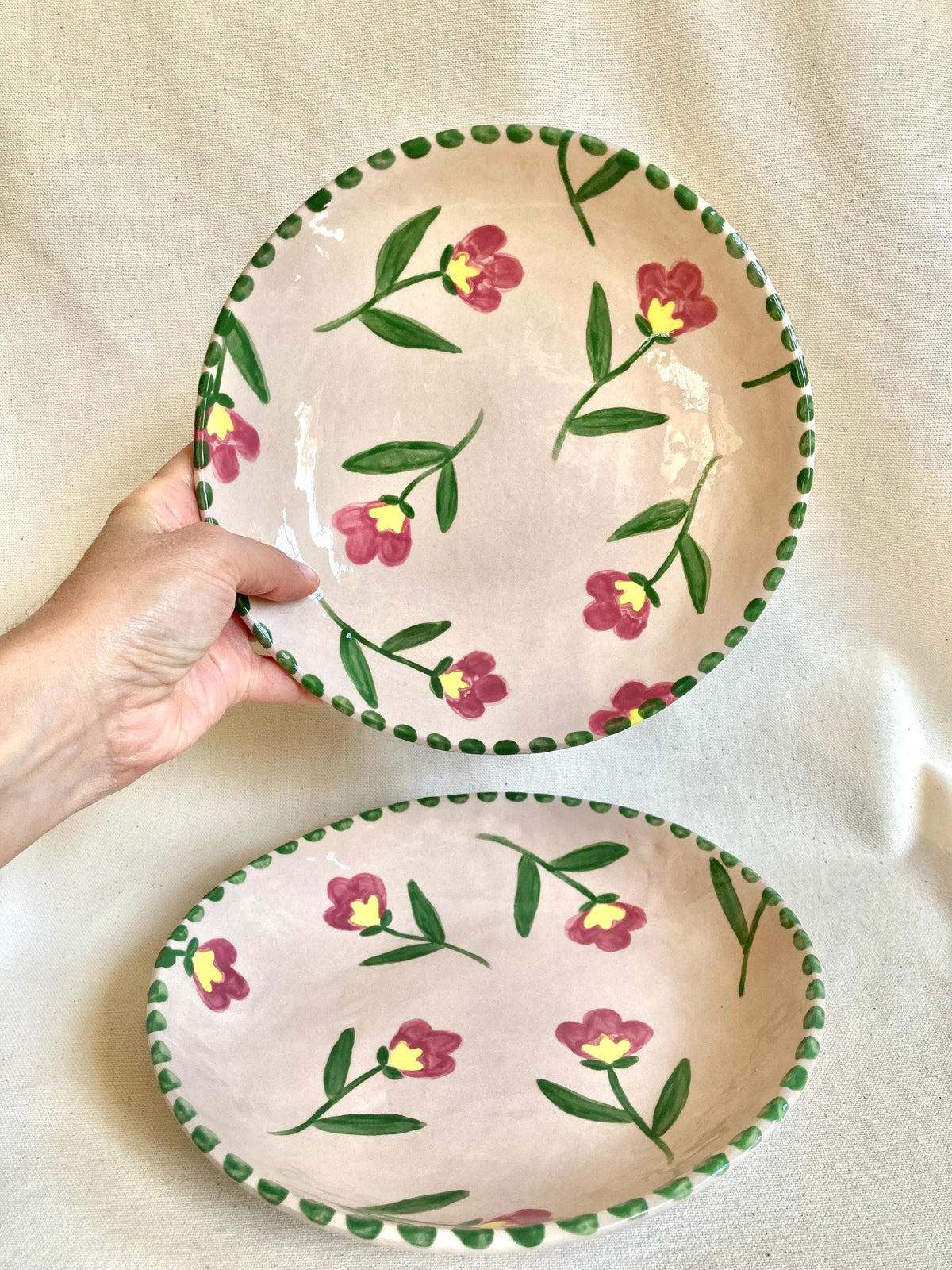 Peut inclure: Deux assiettes en céramique avec un fond rose et un motif floral. Les fleurs sont rouges avec des centres jaunes et des tiges et feuilles vertes. Les bords sont décorés d'une bordure verte pointillée. Une assiette est ronde, l'autre ovale.
